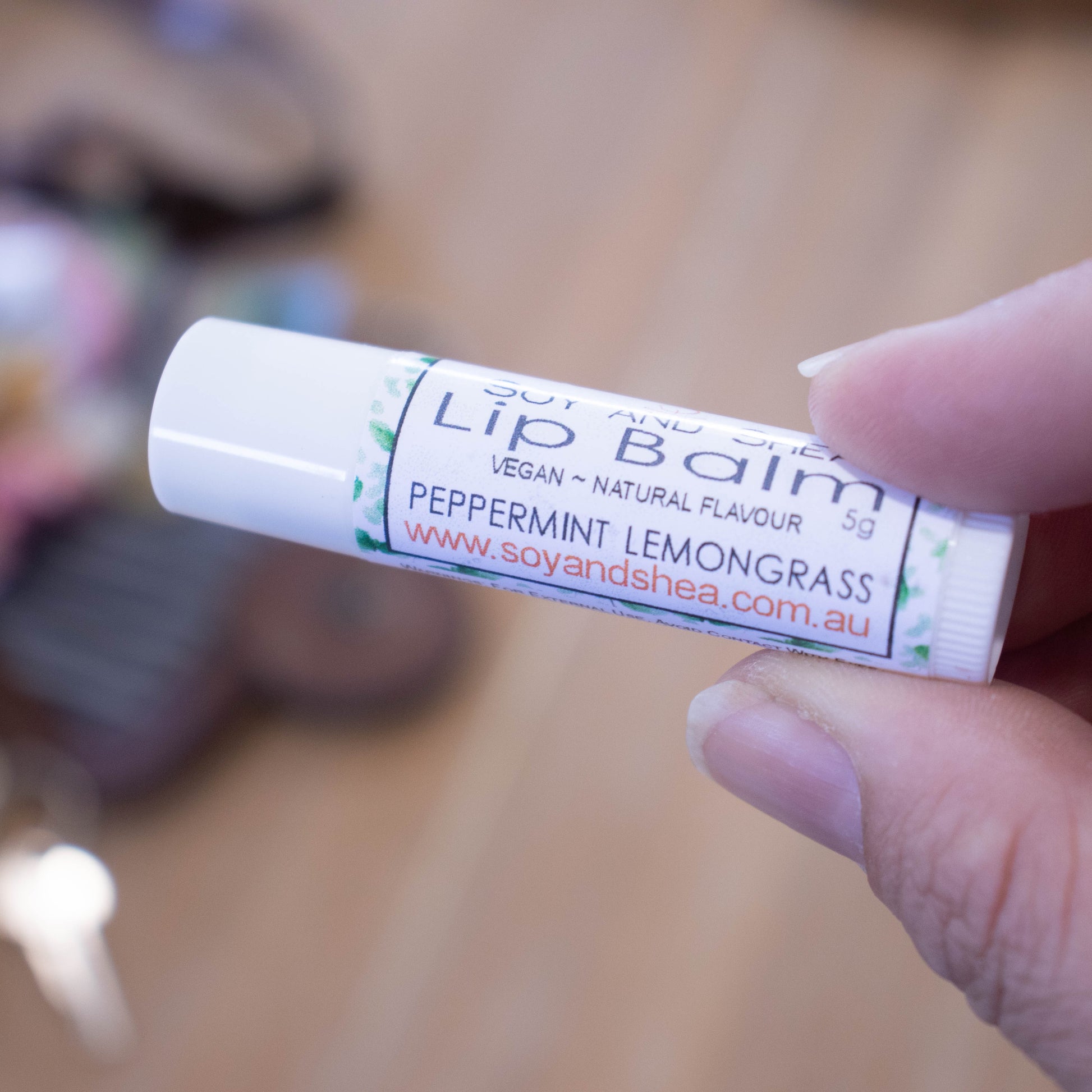 A hand holds a white tube labelled lip balm over a blurred image of an open handbag with contents spilling onto the table.  The lip balms features a white label with a random scattering of green leaves