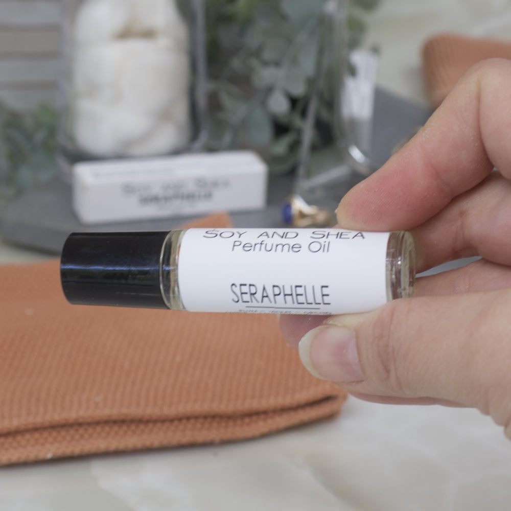 Hand holding a small bottle of Seraphelle perfume oil with a blurred background.  The bottle is clear glass with a black lid.  Around the bottle is a white label