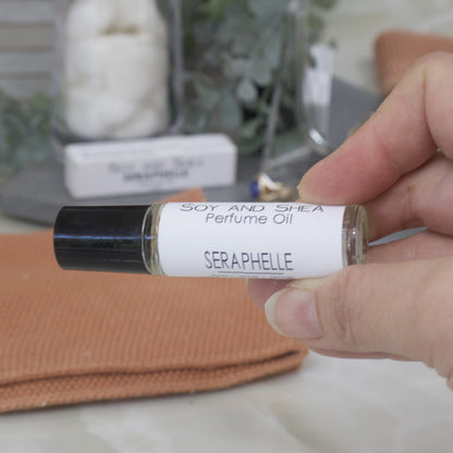 Hand holding a small bottle of Seraphelle perfume oil with a blurred background.  The bottle is clear glass with a black lid.  Around the bottle is a white label