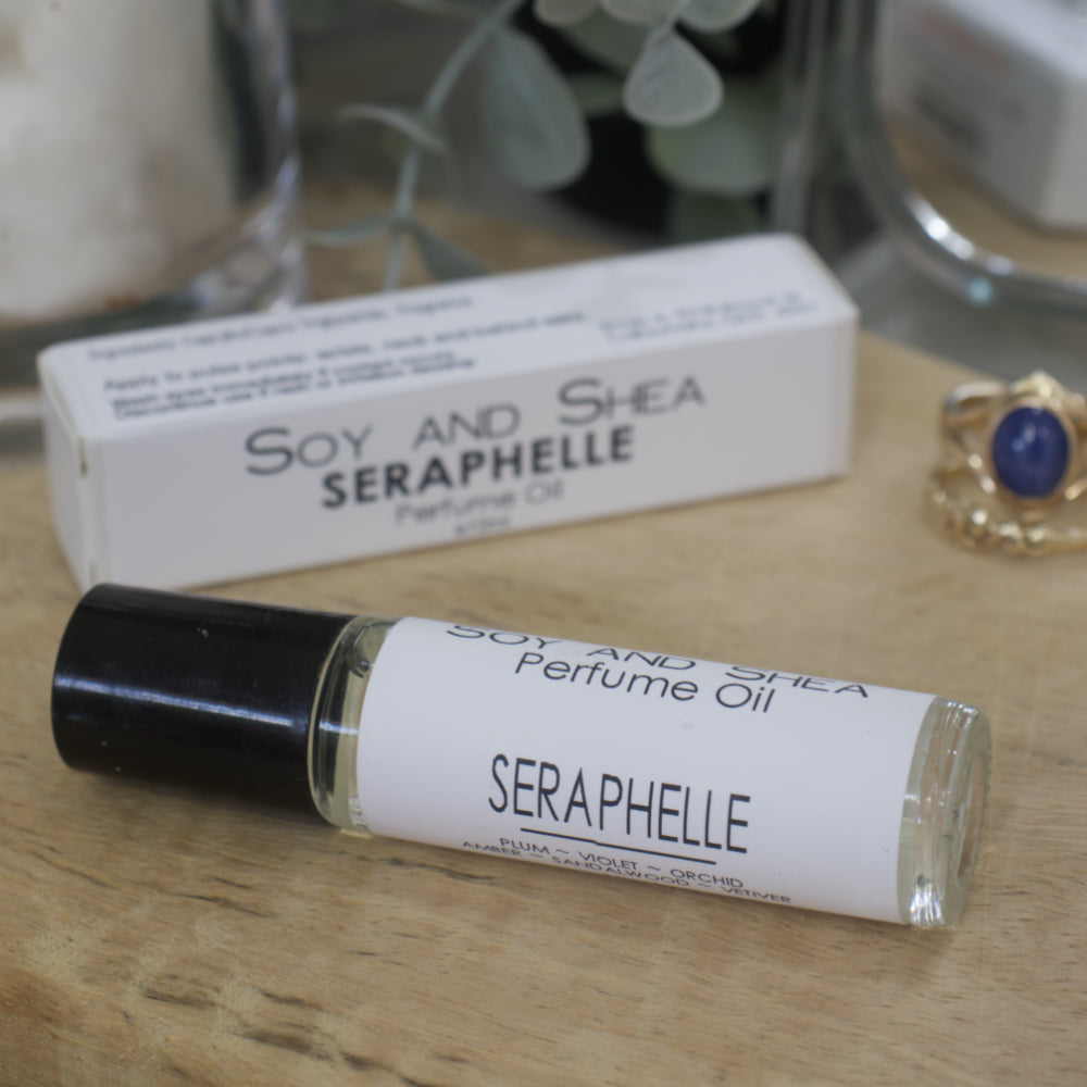 Seraphelle Soy and Shea perfume oil bottle on a wooden surface with packaging in the background.  The bottle is clear glass with a black lid and a wrap around white label