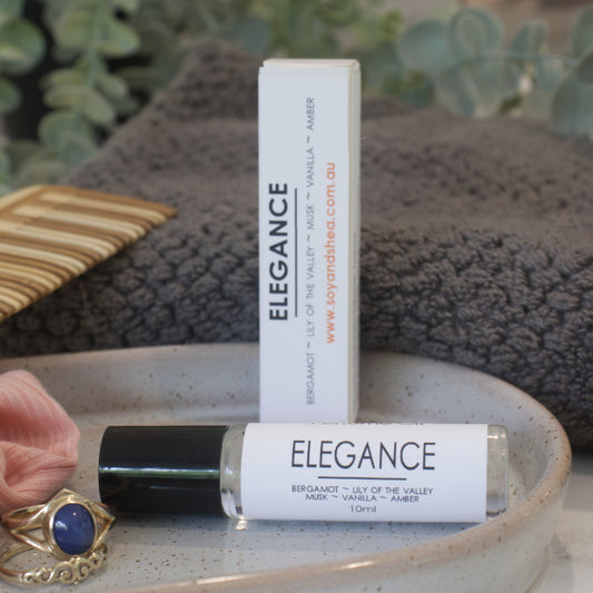A clear glass roller bottle with black lid, of Elegance Perfume Oil with a white label and a box, displayed on a ceramic plate with a grey towel and a piece of jewelry.