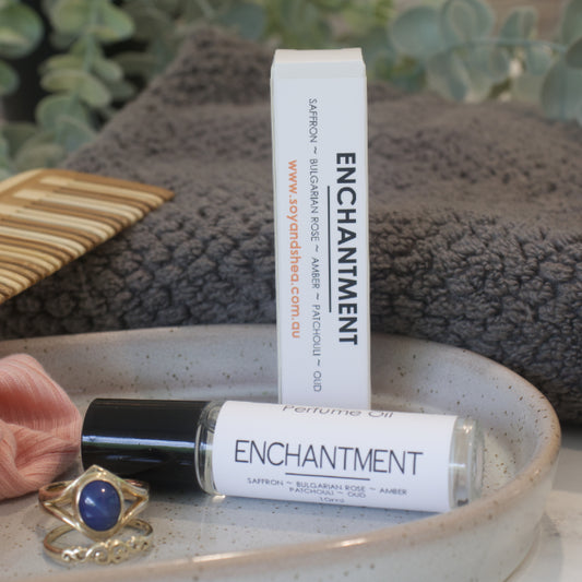 A clear glass roller bottle with black lid,  of Enchantment Perfume Oil with a white label and a box, displayed on a ceramic plate with a grey towel and a piece of jewelry.