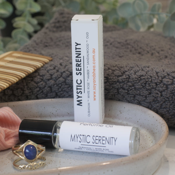 A clear glass roller bottle with black lid,  of Mystic Serenity Perfume Oil with a white label and a box, displayed on a ceramic plate with a grey towel and a piece of jewelry.