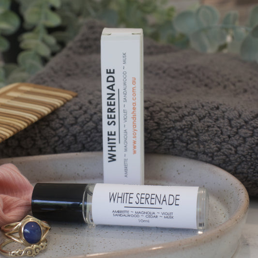 A clear glass roller bottle with black lid,  of White Serenade Perfume Oil with a white label and a box, displayed on a ceramic plate with a grey towel and a piece of jewelry.