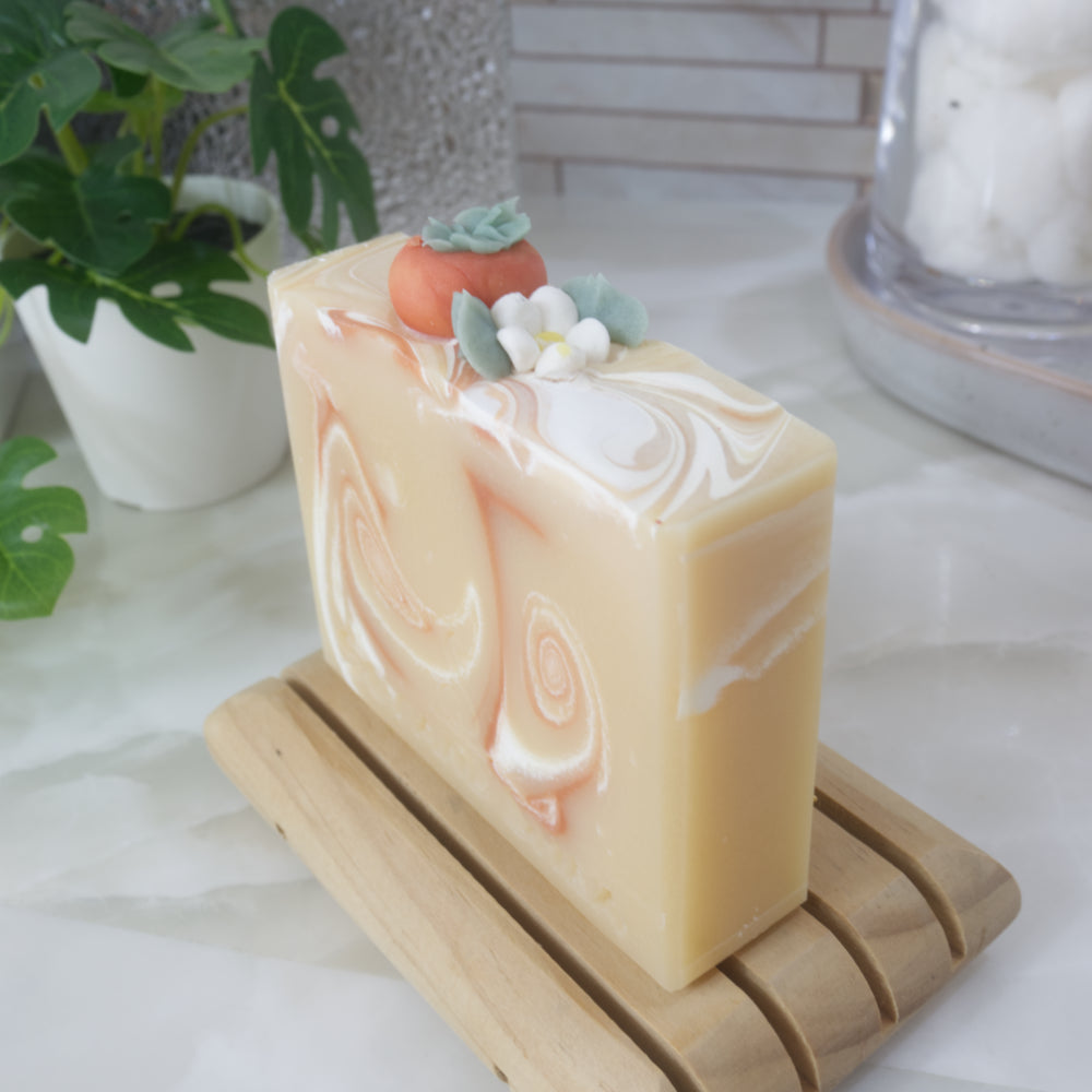 A soap bar stands on a wooden soap rack against a backdrop of a bathroom counter top.  The soap is a pale yellow-orange with dark orange and white swirls throughout.  The top of the soap features a replica of a persimmon fruit along with white flower all made of soap