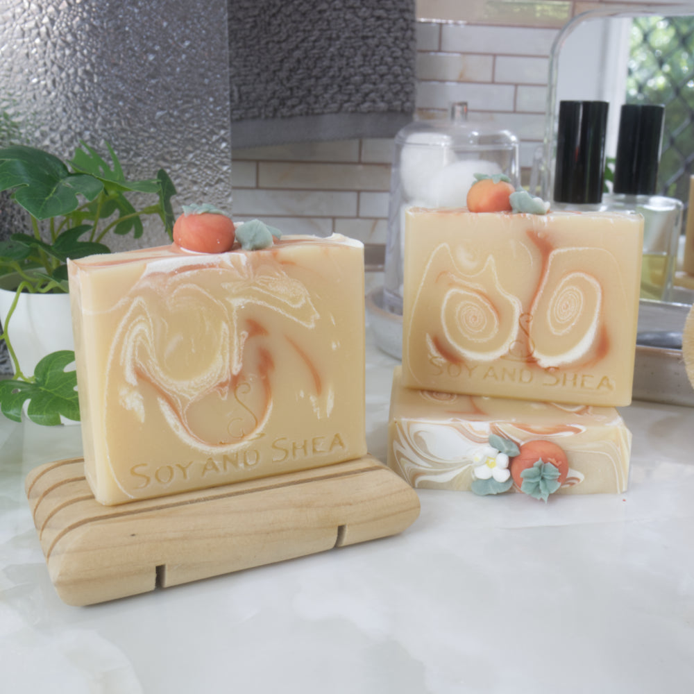 Three decorative soap bars are displayed on a marble bathroom countertop with small mirror, perfume and plant.  One soap lays flat showing the decorations on top, with another soap standing on top.  To the front-left, another soap stands on a wooden soap dish.  All soaps are a pale orange-yellow with varying swirls of white and dark orange.  The soaps tops all feature a replica of a persimmon fruit along with a small white flower made from soap