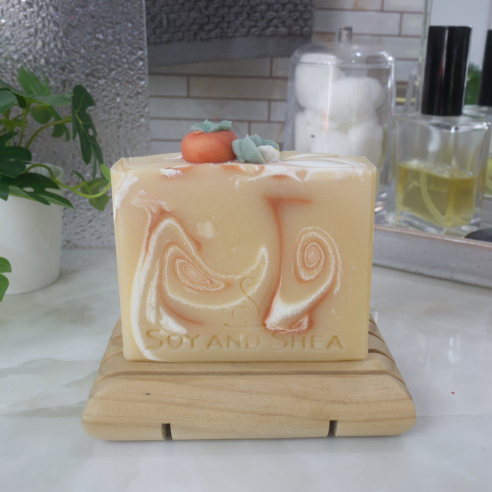 A soap bar stands on a wooden soap rack against a backdrop of a bathroom counter top with perfume, plant and mirror.  The soap is a pale yellow-orange with dark orange and white swirls throughout.  The top of the soap features a replica of a persimmon fruit along with white flower all made of soap