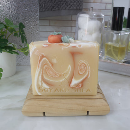 A soap bar stands on a wooden soap rack against a backdrop of a bathroom counter top with perfume, plant and mirror.  The soap is a pale yellow-orange with dark orange and white swirls throughout.  The top of the soap features a replica of a persimmon fruit along with white flower all made of soap