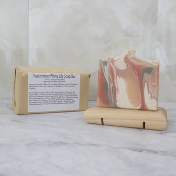 A soap bar with orange, green and white swirls sits on a wooden stand with a brown paper package behind labeled 'Persimmon White Lily Soap Bar' on a marble background