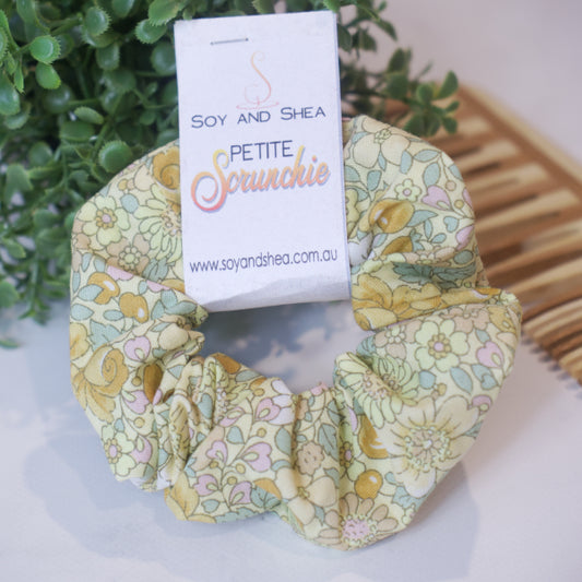 A colorful scrunchie sits on wooden surface resting against a wooden comb with greenery in the background.  A cardboard tag reads SOY AND SHEA PETITE SCRUNCHIE.    The scrunchie features a floral patterm of bramble flowers and berries in yellow and pink tones with pale green leaves against a pale yellow background