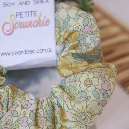 A close up of the fabric design on the Petite Brambles scrunchie depicting bramble flowers in shades of yellow against a pale yellow background along with small leaves.  A cardboard tag is attached to the scrunchie