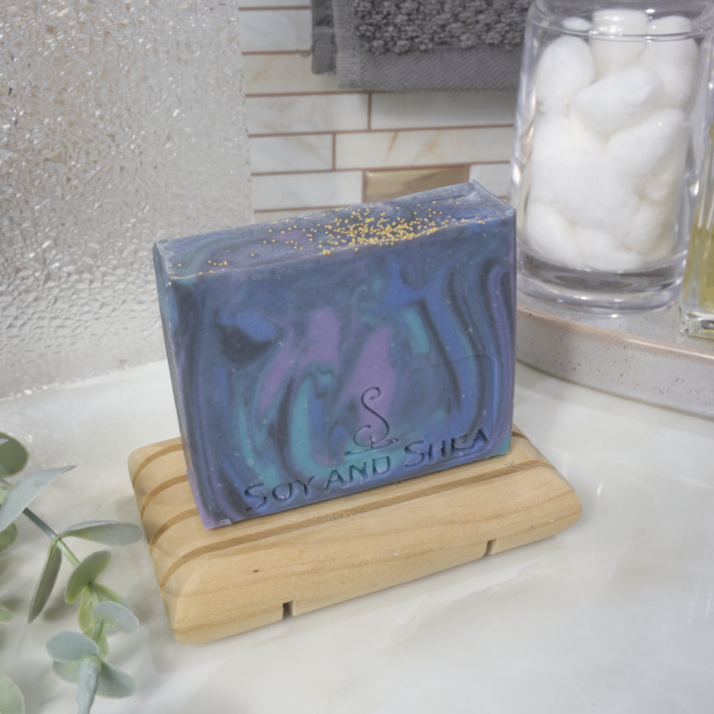  A rectangular bar of soap stands on a wooden soap rack on a bathroom countertop next to a jar of cotton balls and some greenery. The soap has a swirling pattern in shades of blue, black and purple.  The top of the soap is flat and features tiny gold balls on the top.