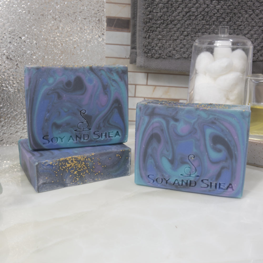 Three rectangular bars of soap sit on a bathroom counter top. One bar lays flat on the bench revealing a flat top with micro gold beads on the top.  A soap stands upright on this bar with another soap standing on the bench to the front right.  All soaps have a swirling colourful pattern in shades of blue, purple and black