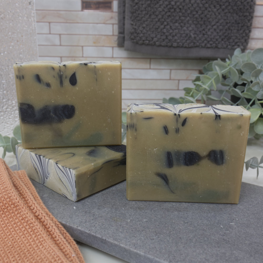 Three bars of pale green soap with black drop patterns on a stone surface with a plant in the background