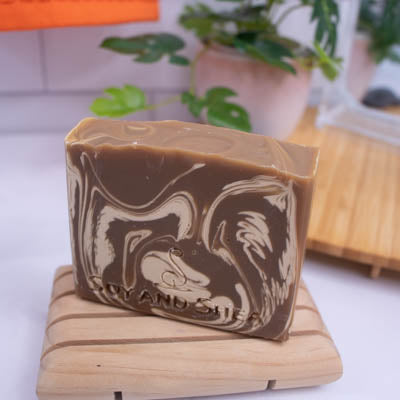 A soap bar stands on a wooden soap dish on a bathroom counter top.  A plant, mirror and towel can be seen in the blured background.  The soap is a rich brown colour with cream swirls.