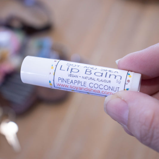 A hand holds a white tube labelled lip balm over a blurred image of an open handbag with contents spilling onto the table.  The lip balms features a white label with illustrations of tiny pineapples and coconuts