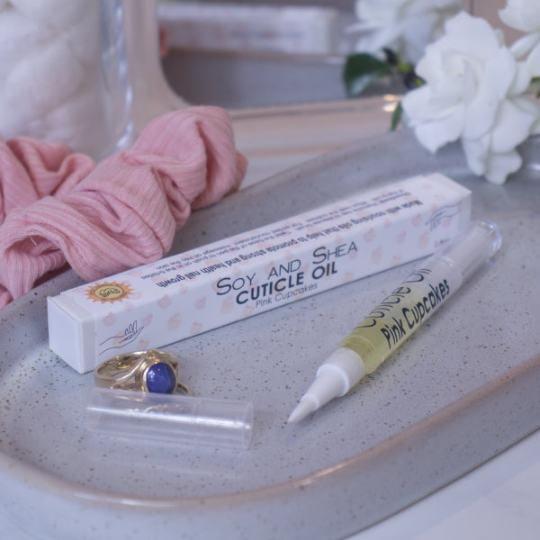 A clear, narrow tube with the lid removed showing a brush tip lies on a stone tray beside a cardboard box labelled CUTICLE OIL and a set of gold rings.  In the background is a bathroom counter top with a pink scrunchie, small mirror, and a some white  flowers.