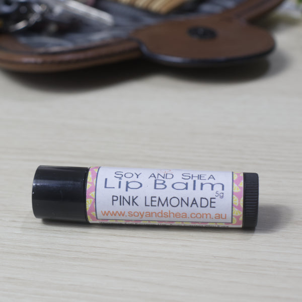 A black tube of Lip balm with 'Pink Lemonade' flavour on a wooden side with blurred image of handback in background.