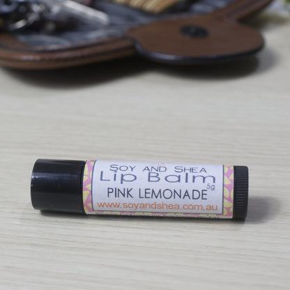 A black tube of Lip balm with 'Pink Lemonade' flavour on a wooden side with blurred image of handback in background.