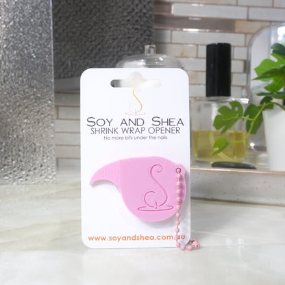 A light pink Shrinkwrap opener with matching ball and chain keyring is attached to a white card with product details.  The card is standing upright on a bathroom countertop with a bottle of perfume, a jar of cotton balls and towel in the background