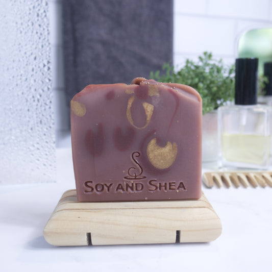 A bar of soap on a wooden dish, with 'SOY AND SHEA' branding, placed on a countertop in a bathroom setting.  The soap has a dusky red colour with deep red and and gold drops.