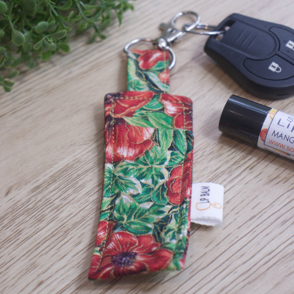 A Lip Balm holder lies on an wooden surface with lobster claw clasp attached to a car key.  A lip balm and greenery can be seen in the image.  The Clip Balm has a bold print of large red poppies with purple centres surround by green leaves.