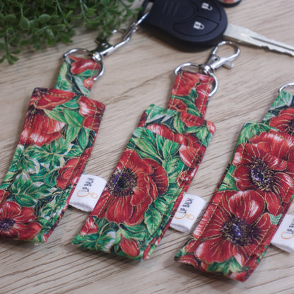 A selection of three lip balms showing variations in fabric design, lie on a wooden side with one attached to a car key with a lobster claw clasp.  The Clip balms have a bold pattern of large red poppies with purple/black centres surrounded by bright green leaves.