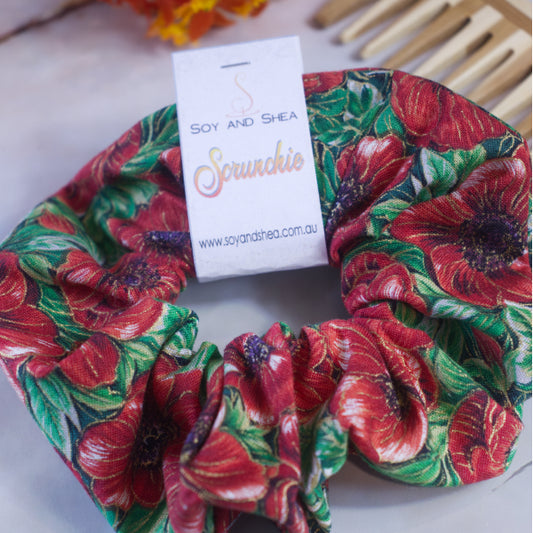 A colorful scrunchie sits on wooden surface resting against a wooden comb with greenery in the background.  A cardboard tag reads SOY AND SHEA SCRUNCHIE.  The Scrunchie features a bold pattern of large red poppies with purple centres and gold highlights against a bed of bright green leaves