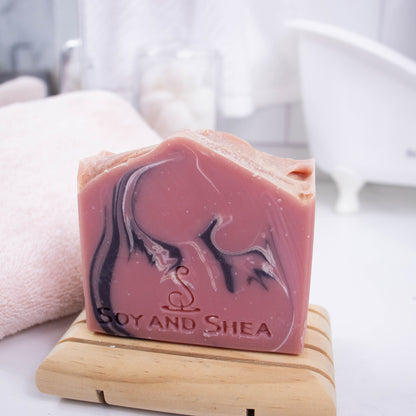 A dusky pink soap bar with black and white swirls, stands on a wooden soap dish along side a white towel with a blurred bathroom counter top