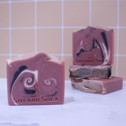 A collection of soaps sit on a white marble surface against a pale peach tile background.  In the background, two soaps lay flat but stacked upon each other revealing a wavy textured top.   A third soap stands on stop of this stack with a fourth soap in the foreground.  The soaps are a rich dusky pink with white and black curling swirls.  Each soap shows a slight variation in design.