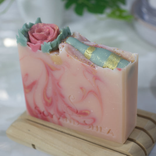 A close up image of a soap bar resting on a wooden soap rack with a blurred background.  The soap is pale pink with swirls of dark pink and gold.  The top of the soap features a single piped red rose with green leaves to one side of the soap with the other featuring a green wine bottle with gold details.  