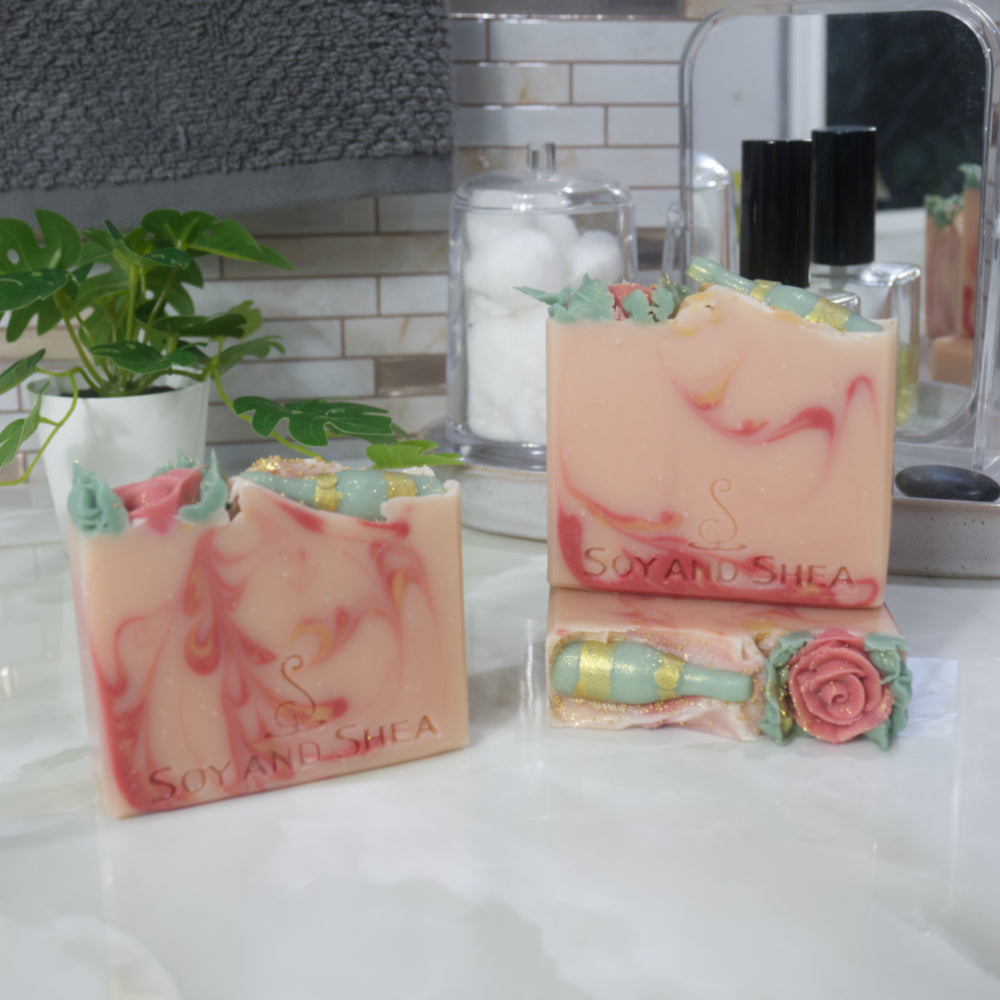 A select of three soap are displayed on marble bathroom countertop with a plant, perfume bottle and mirror.   One soap lays flat showing the top of the soap which is decorated with a single red rose and a green wine bottle all made with soap.  A soap stands upright on this soap with another to the left of these these.  All soaps are a pale pink with dark pink and gold swirls of varying designs.  All feature the decorative top