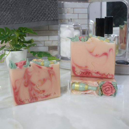 A select of three soap are displayed on marble bathroom countertop with a plant, perfume bottle and mirror.   One soap lays flat showing the top of the soap which is decorated with a single red rose and a green wine bottle all made with soap.  A soap stands upright on this soap with another to the left of these these.  All soaps are a pale pink with dark pink and gold swirls of varying designs.  All feature the decorative top