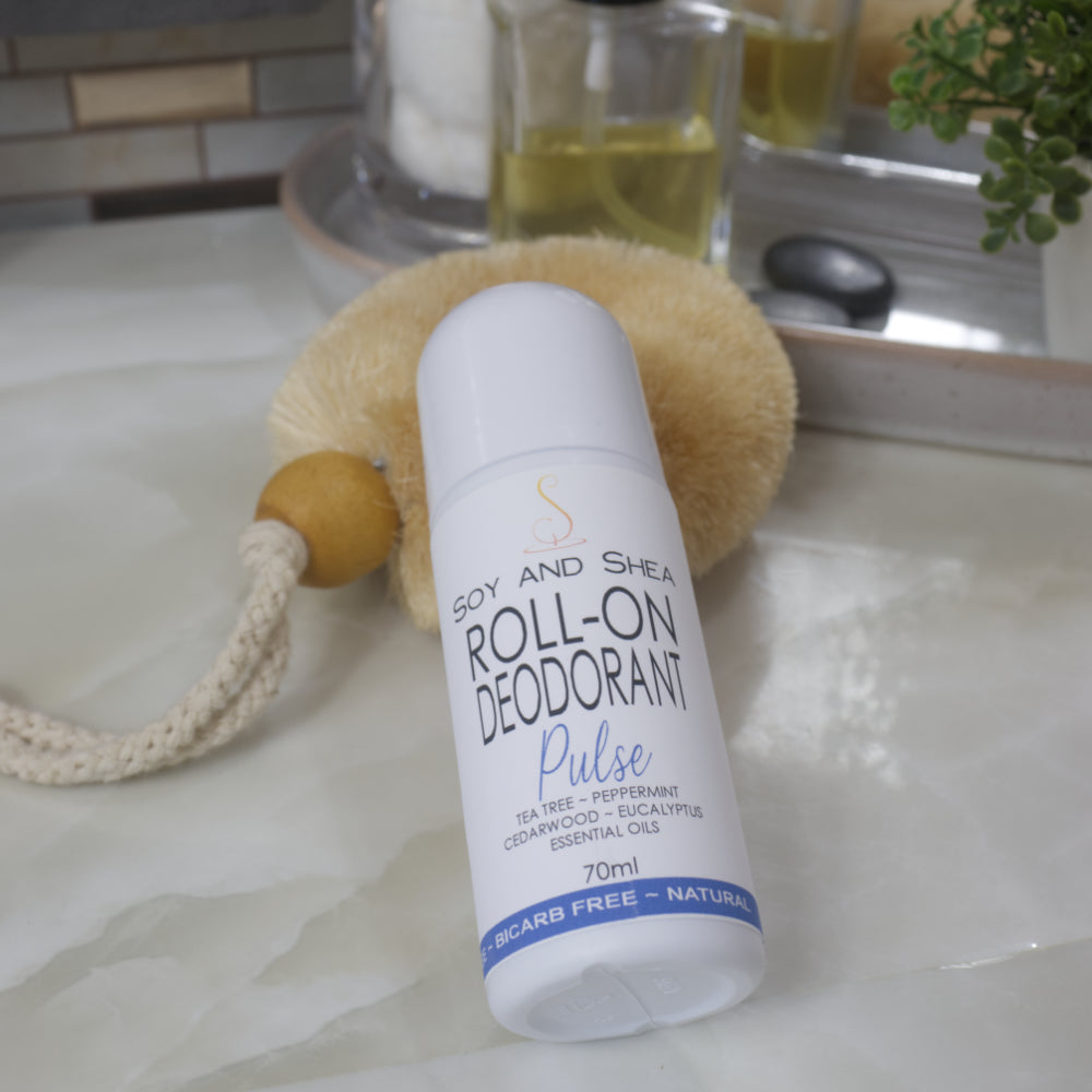 A white bottle with domed lid lays resting against a natural body brush on a bathroom countertop.  The product label identifies it as Pulse Roll-on Deodorant with a list of essential oils 