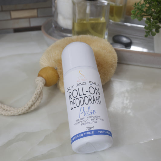 A white bottle with domed lid lays resting against a natural body brush on a bathroom countertop.  The product label identifies it as Pulse Roll-on Deodorant with a list of essential oils 