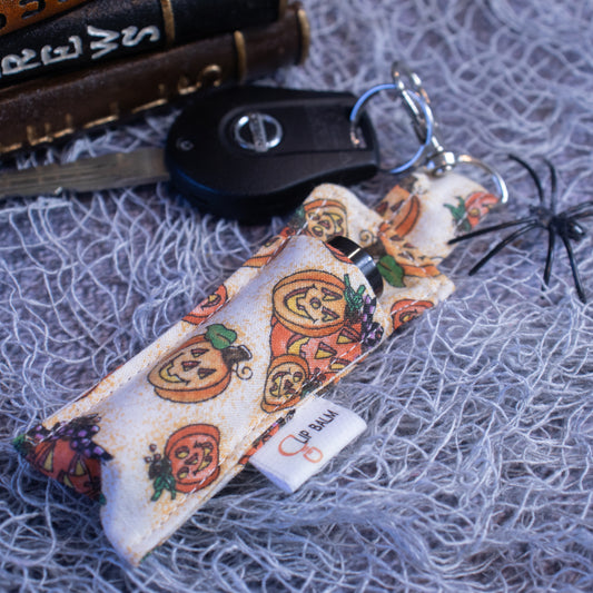 A Lip Balm holder lies on a grey mesh fabric attached to a car key with a lobster claw clasp.  The Clip balm is cream in colour with illustrations of orange glowing Jack-o-Lanterns in various sizes