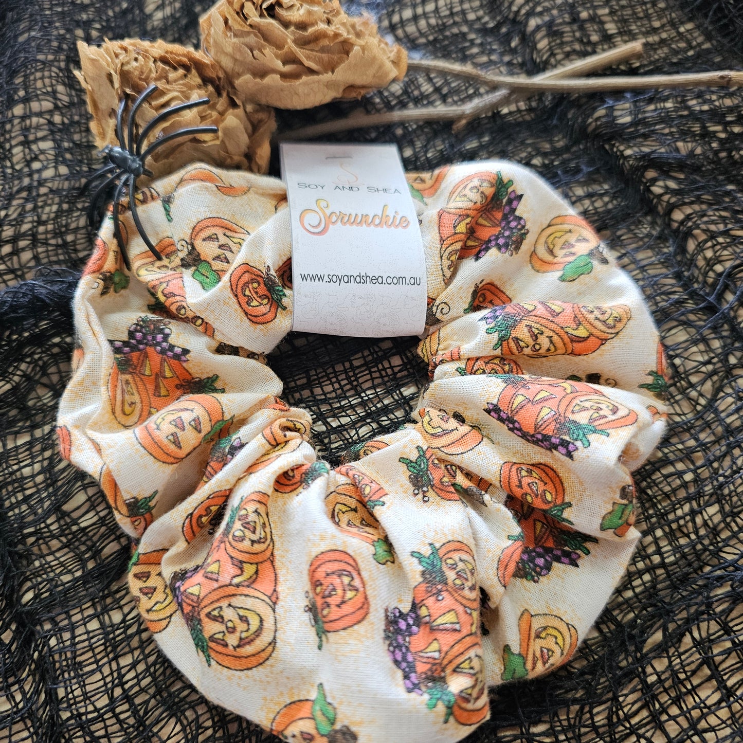A colorful scrunchie sits on wooden surface covered in a black mesh fabric with dried roses in the background.  A cardboard tag reads SOY AND SHEA SCRUNCHIE.  The Scrunchie is cream with illustrations of glowing orange jack-o-lanterns of various sizes 