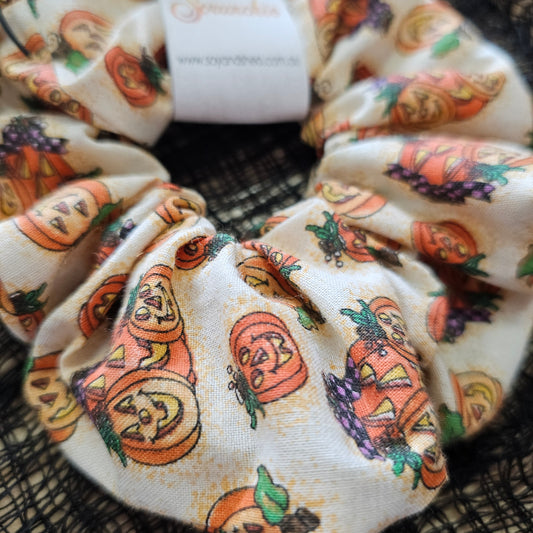 A detailed view of the Pumpkin Patche Scrunchie showing a cream fabric covered in illustrations of orange, glowing jack-o-lanterns in various sizes