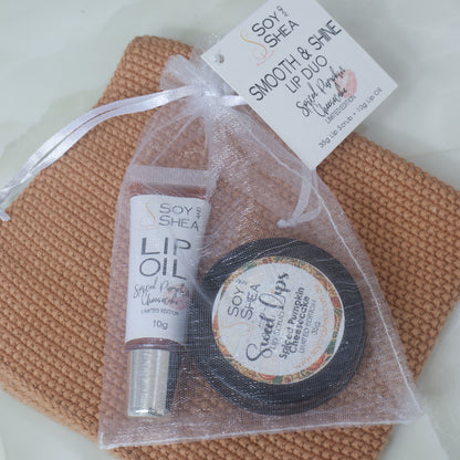 A black tin of Sweet Lips Lip Scrub and a tube of Lip oil are inside a white organza bag resting on an orange face cloth.  The bag is labelled as Smooth & Shine Lip Duo in a Spiced Pumpkin Cheesecake flavour