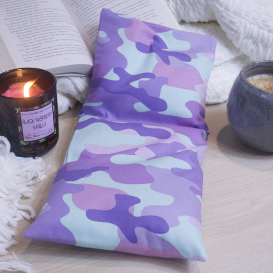 A rectangular heat pack with a purple camouflage pattern, placed on a wooden surface, with a lit candle and an open book in the background.
