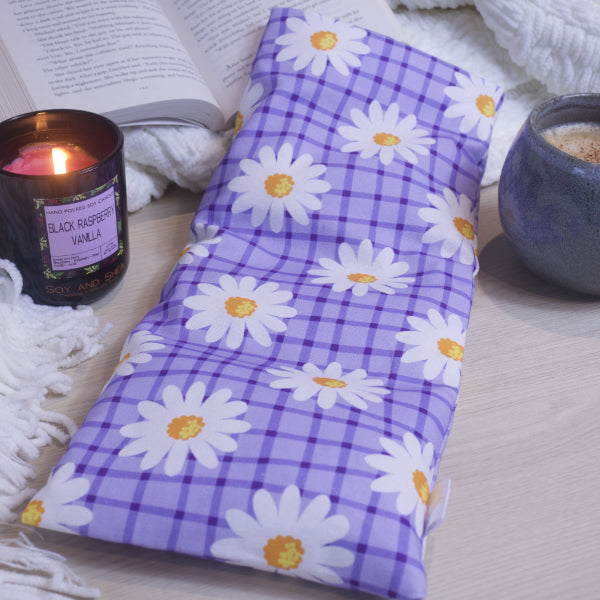 A rectangular heat pack with a purple checkered pattern and white daisy designs, displayed on a table alongside a lit candle and an open book.