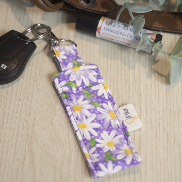 A clip balm holder lies on a wooden surface attached to a car key with a "lobster claw" clip.  To the side is a tube of lip balm surrounded by greenery and a leather strap from a bag.  The Clip Balm has a purple background with a arrany of white daisies with yellow centres and green leaves scattered over. 