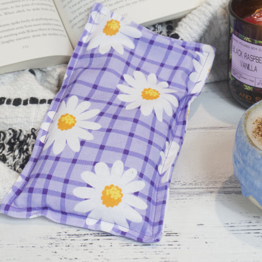 The Soy and Shea Purple Daisies Mini Heat Pack rests beside a book and candle, creating a cosy scene.   The small rectangular heat pack has a light puprle background with dark purple checkered pattern over it.  White daisies with yellow centres are scattered over it.