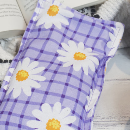 A close up photo of the Purple Daisy Mini Heat pack showing the fabric design of a Purple checkered background with white daisies with yellow centres scattered over.  The edge of the heat pack rests against a book and black and white blanket.