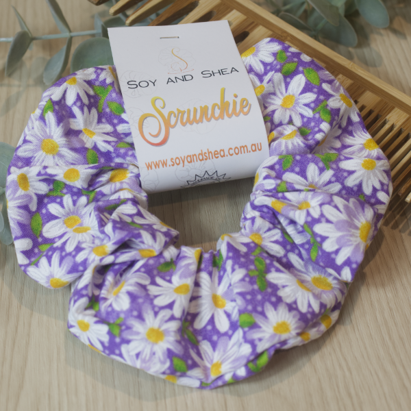 A colorful scrunchie sits on wooden surface resting against a wooden comb with greenery in the background.  A cardboard tag reads SOY AND SHEA SCRUNCHIE.    The scrunchie has bright purple background with illustrations of daisies with white centers scattered throughout