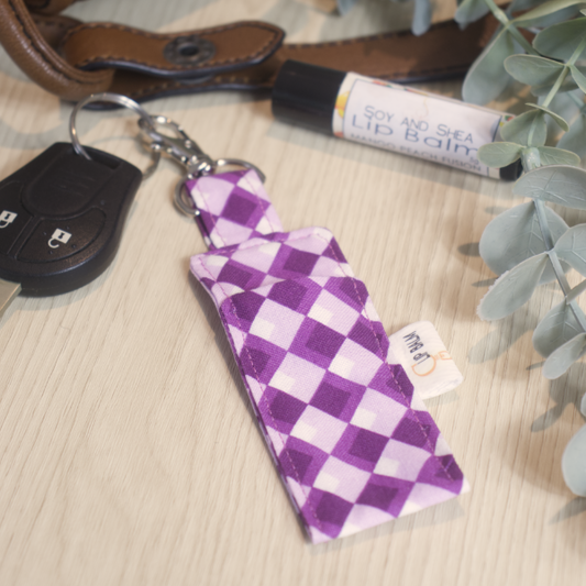 A clip balm holder lies on a wooden surface attached to a car key with a "lobster claw" clip.  To the side is a tube of lip balm surrounded by greenery and a leather strap from a bag.  The Clip Balm features a harlequin, diamond design running up the length in shades of purple