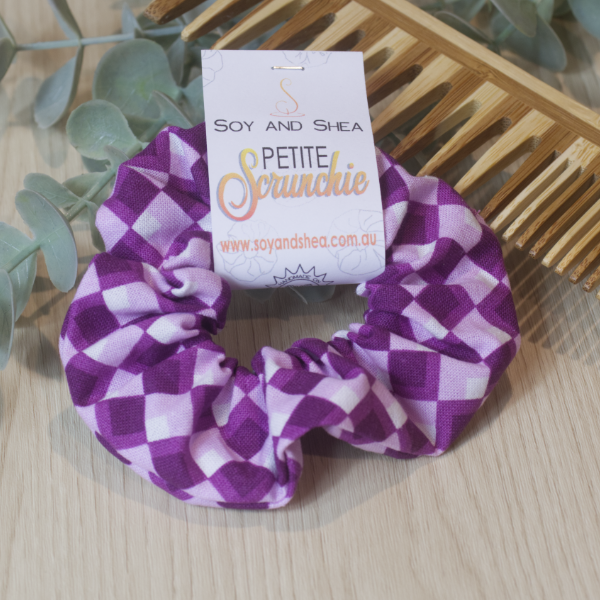 A purple and white harlequin patterned petite scrunchie, displayed resting on a wooden comb with a Soy and Shea packaging behind it.