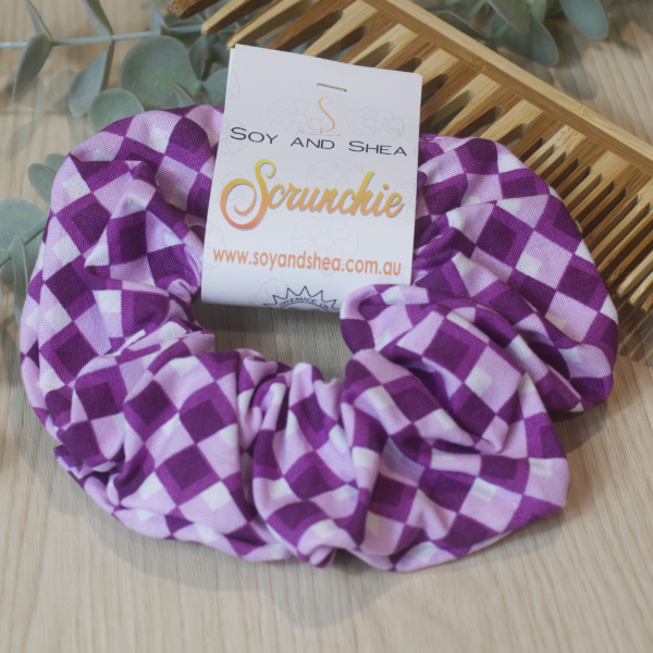 A colorful scrunchie sits on wooden surface resting against a wooden comb with greenery in the background.  A cardboard tag reads SOY AND SHEA SCRUNCHIE.    The design has a harlequin style diamond pattern in shades of purple and white