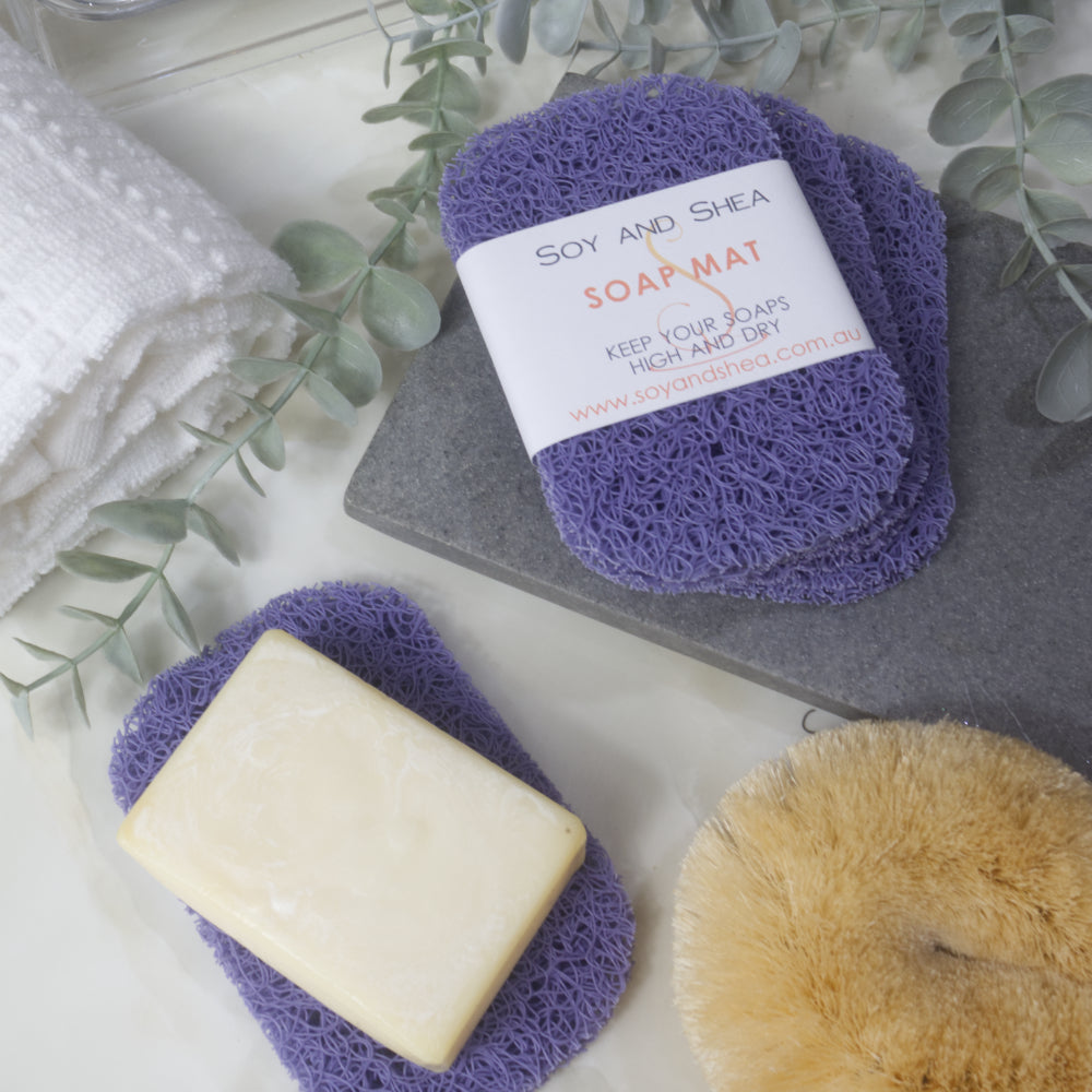 A flatlay image showing a collection of Purple Soap mats, displayed on a bathroom countertop along with a white towel, body brush and greenery.  Three soap mats are stacked upon each other in a fan shape on a grey stone tray with the top one featuring a product label.  A fourth mat lies to the bottom of the image with a cream soap lying on it.
