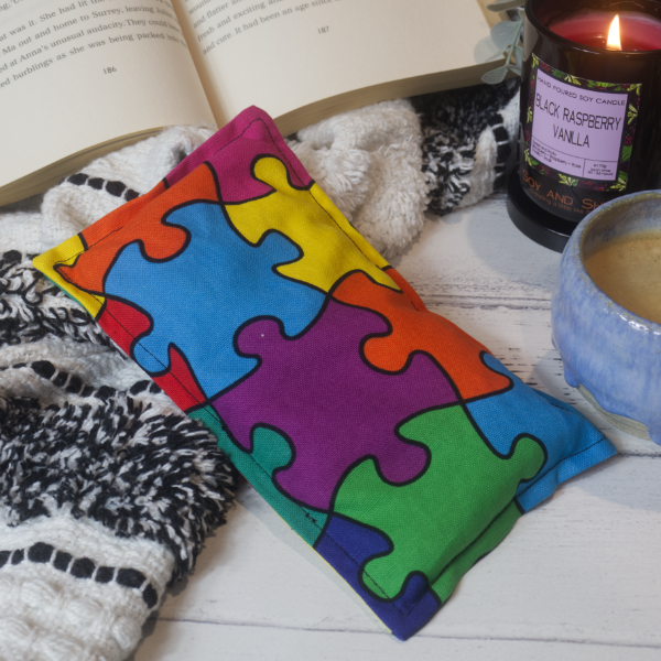 A mini rectangular heat pack rests against a black and white blanket and open book.  Next to it is a burning candle and filled coffee cup evoking a comfy scene.   The Heat Pack features a design of bright coloured puzzle pieces all connected